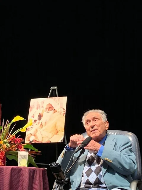Ishwar Puri speaking while seated beside a display of flowers