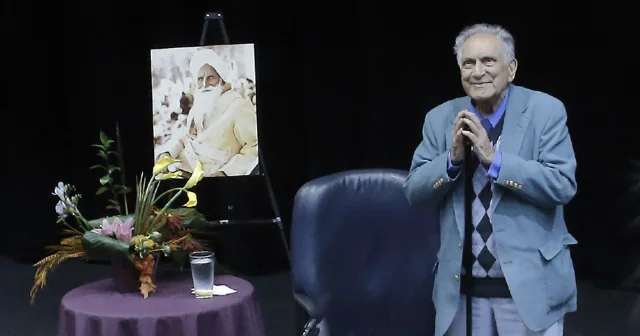 Ishwar Puri addressing an audience from a stage, wearing a blue jacket